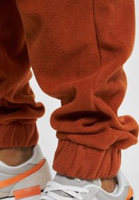 Rust orange fleece pants with gathered cuffs, paired with light gray sneakers featuring white and orange accents. Smooth texture and relaxed fit.