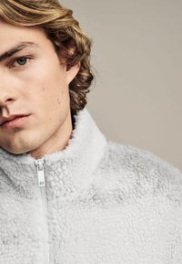 Jasnoszara polarowa kurtka z wysokim kołnierzem i pełnym zamkiem błyskawicznym. Charakteryzuje się miękką, fakturowaną powierzchnią o puszystym wyglądzie.