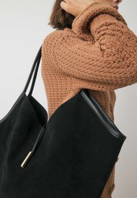 Melnas zamšādas somiņa ar ādas akcentiem, kura izceļas ar elegantu, leņķainu formu un zeltītiem elementiem. Redzama mīksta tekstūra.