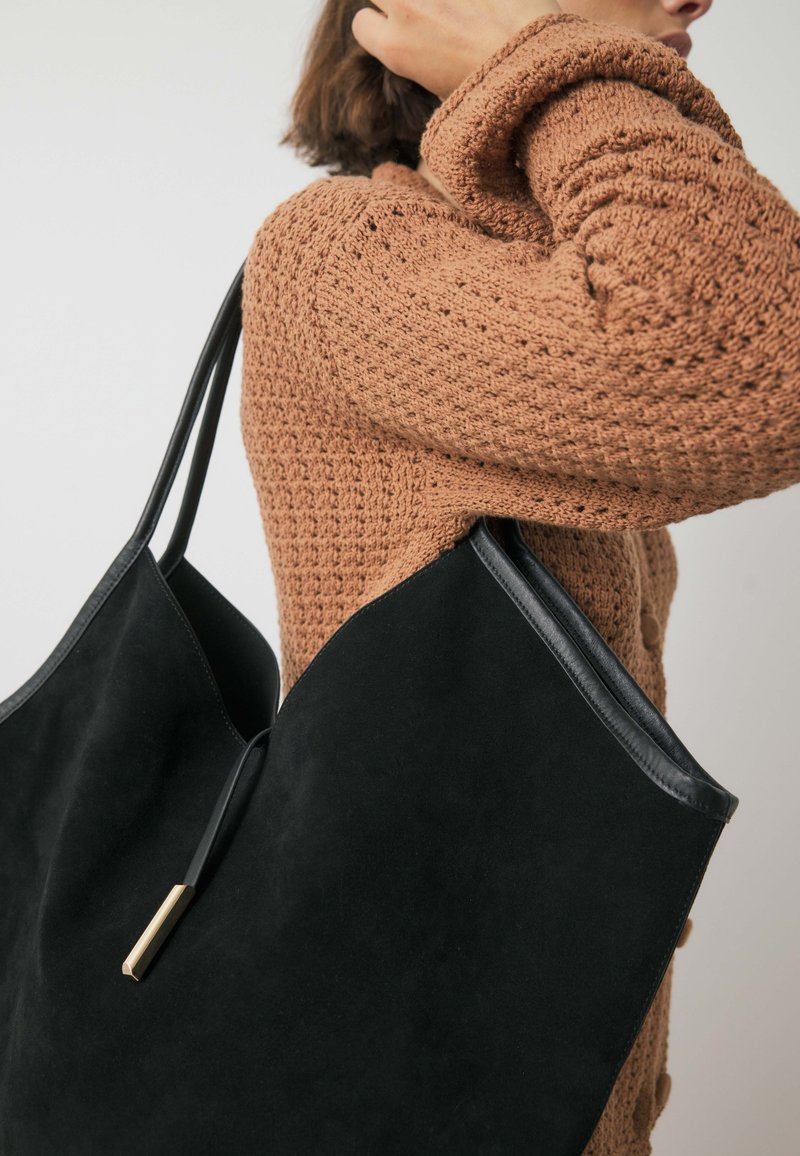 Melnas zamšādas somiņa ar ādas akcentiem, kura izceļas ar elegantu, leņķainu formu un zeltītiem elementiem. Redzama mīksta tekstūra.