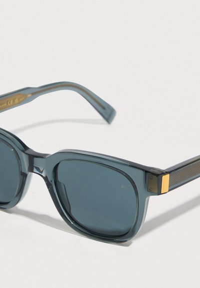 Gafas de sol cuadradas translúcidas azules con lentes oscuras y detalles dorados en las patillas, descansando sobre una superficie pálida.