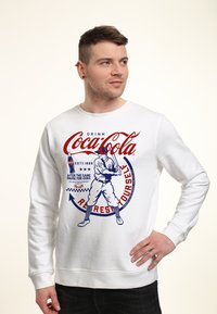 Weißer Sweatshirt mit einem Vintage Coca-Cola Grafik, die einen Baseballspieler und roten-blauen Text enthält, mit gerippten Bündchen und Saum.