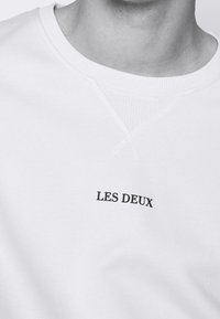 White sweatshirt featuring a round neckline and ribbed cuffs. The brand name "LES DEUX" is printed in black at the center. Soft texture.