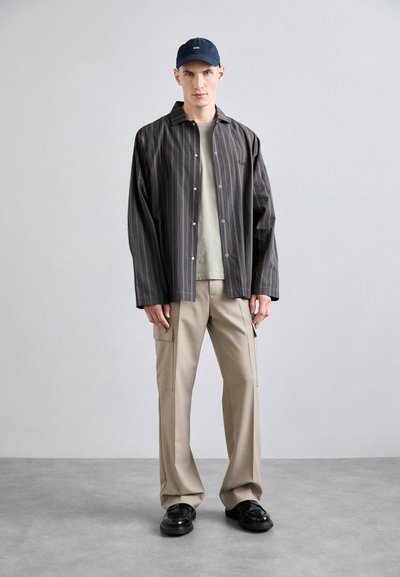 Chemise à boutons en tissu rayé foncé, pantalon beige avec poches cargo, et chaussures noires. Complétée par une casquette marine. Design simple et moderne.
