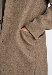 Manteau en laine marron au toucher doux, avec une fermeture à un bouton et une poche latérale. Comprend des boutons noirs au niveau des poignets.