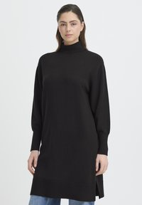 Fekete kötött tunika magas nyakkal, hosszú ujjakkal, oldalsó hasításokkal és bordázott szegéllyel, világoskék farmerral párosítva.