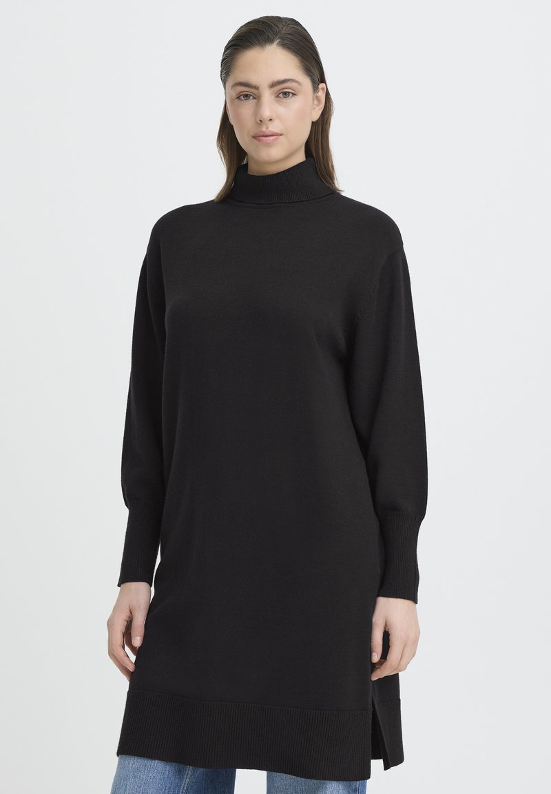 Fekete kötött tunika magas nyakkal, hosszú ujjakkal, oldalsó hasításokkal és bordázott szegéllyel, világoskék farmerral párosítva.