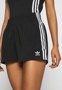 Černé sportovní šortky s elastickým pasem, na bocích s bílými pruhy a vyšitým logem Adidas v dolním levém rohu.