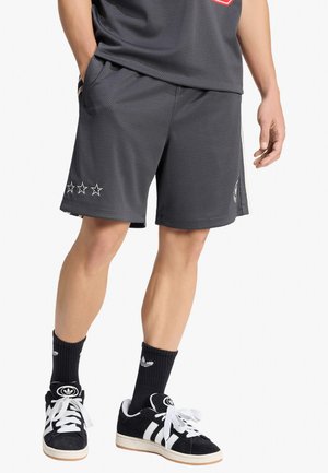 Parte inferiore del corpo di una persona che indossa pantaloncini a rete neri con tre stelle, calze nere Adidas e scarpe da ginnastica Adidas nere e bianche.