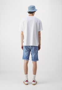 Bílá bavlněná tričko, světle modré džíny šortky, bílé ponožky a tenisky s červenými akcenty. Modrý klobouk s vzorem. Neformální oblečení.