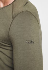 Cămașă lungă cu mânecă, de culoare verde măslinie, realizată dintr-un material texturat, cu o croială slim și un mic logo brodat pe brațul superior.