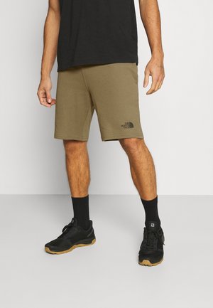 Homme portant un short olive The North Face, des chaussettes noires, des chaussures de sport noires et un t-shirt noir, se tenant devant un fond uni.