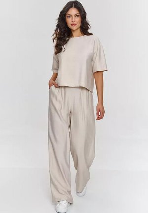 Kobieta z długimi kręconymi włosami, ubrana w luźny beżowy top i szerokie spodnie, idzie do przodu z jedną ręką w kieszeni, białe trampki.