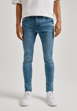 Homme portant un jean slim bleu déchiré et des baskets blanches, debout devant un fond clair uni.