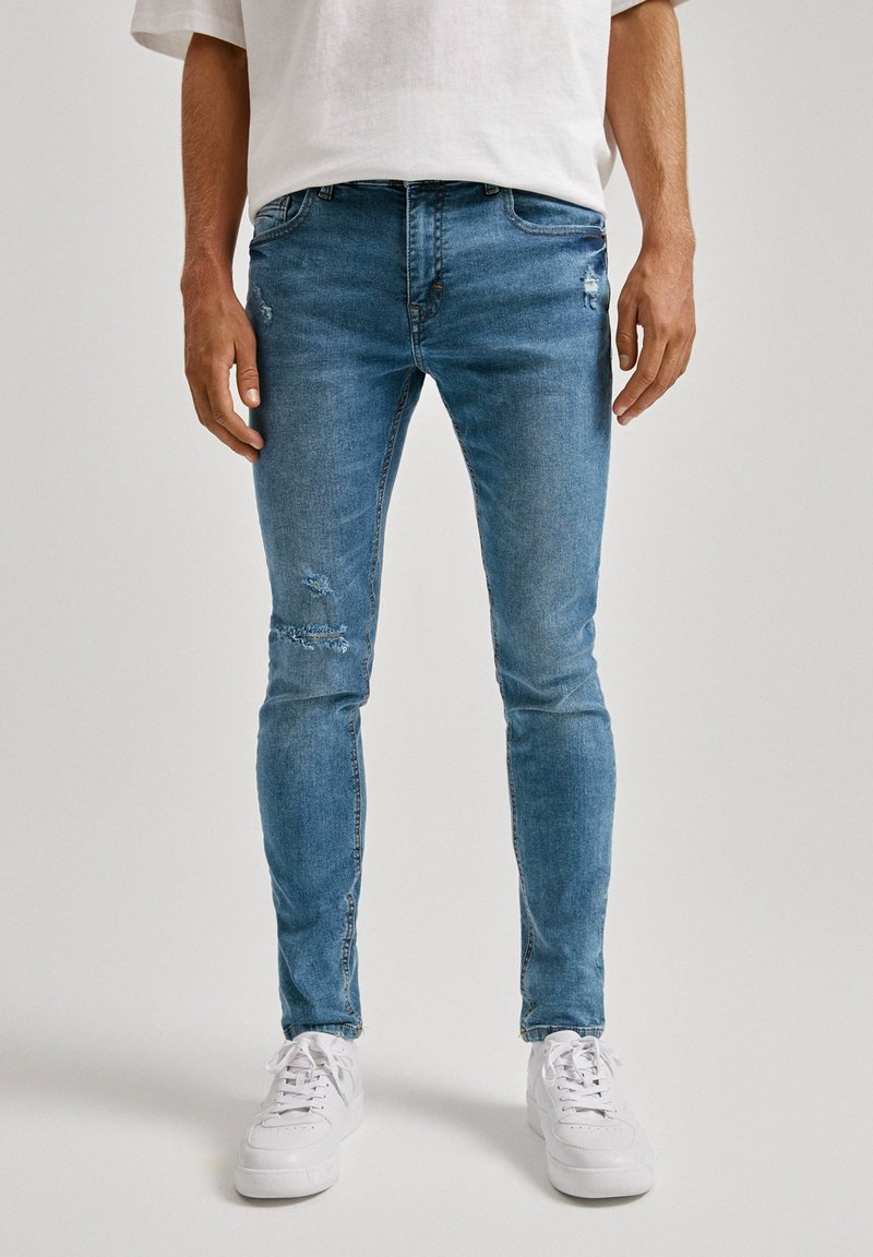 Homme portant un jean slim bleu déchiré et des baskets blanches, debout devant un fond clair uni.
