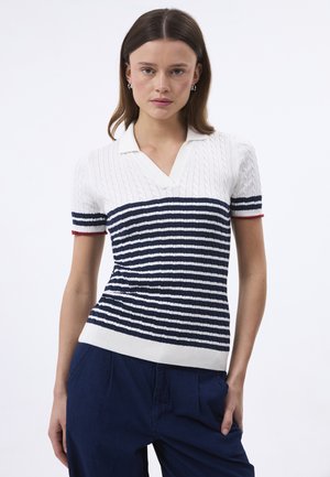 Frau trägt ein weißes und marineblau gestreiftes kurzärmliges Stricktop mit Kragen und marineblaue Hose, steht vor einfachem Hintergrund.