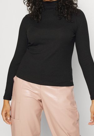 Vrouw draagt een zwarte, geribbelde coltrui met lange mouwen en lichtroze, hooggetailleerde broek met zakken, staand tegen een effen achtergrond.