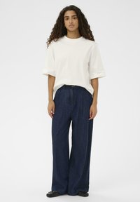 Ženska s kodrasto pričesko, oblečena v ohlapno belo majico, temne modre hlače širokega kroja z visokim pasom in črnimi balerinkami, stoji ob belem ozadju.