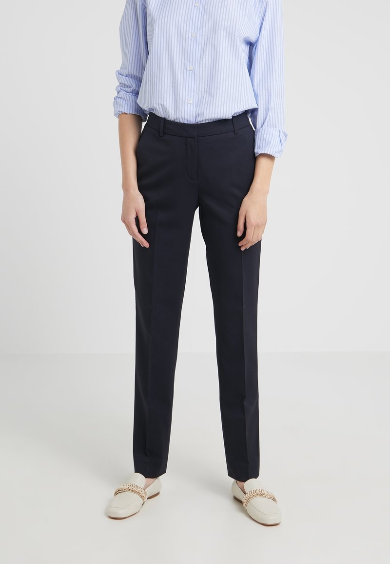 Personne portant un pantalon noir sur mesure, une chemise rayée bleu clair à manches retroussées et des mocassins blancs avec un détail de chaîne en or.