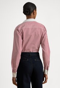 Γυναίκα με σκούρα μαλλιά σε κότσο, που φοράει κόκκινο και άσπρο πουκάμισο με κάθετες ρίγες και λευκό γιακά, καθώς και σκούρο παντελόνι ψηλόμεσο, που κοιτάζει προς τα πίσω.