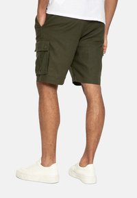 Homme portant un short cargo vert olive et des baskets blanches, debout les mains dans les poches, tourné de dos sur un fond uni.