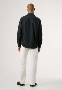 Dunkelblauses Hemd mit langen Ärmeln, geknöpften Manschetten und spitzem Kragen, kombiniert mit weißen Jeans und dunklen Schuhen vor einem schlichten Hintergrund.