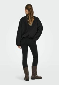 Chemise noire oversized à manches longues avec un col rond ; associée à un jean skinny noir. Le mannequin porte des bottes en cuir marron montant jusqu'aux genoux.
