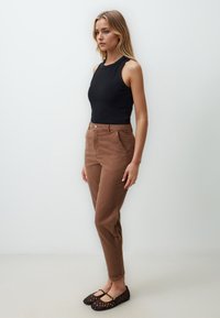 Chinos marroni con tasche anteriori, zip e chiusura a bottone. Abbinati a una canotta nera e a delle scarpe flats nere intrecciate, su uno sfondo neutro.