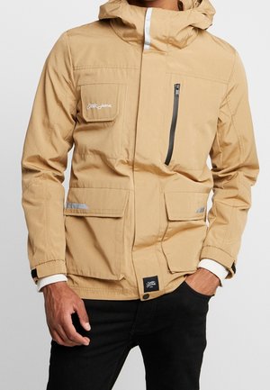 Veste imperméable beige avec capuche, poche poitrine zippée, deux poches avant et accents réfléchissants. Texture lisse, poignets ajustables.