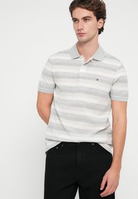 Camisa polo a rayas gris y blanca con un cuello tipo polo y tres botones, combinada con jeans negros. Presenta un logo en el pecho.