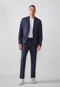 Costume à carreaux bleu marine avec une veste ajustée et un pantalon sur mesure, associé à un t-shirt blanc et des baskets blanches, texture de tissu lisse.