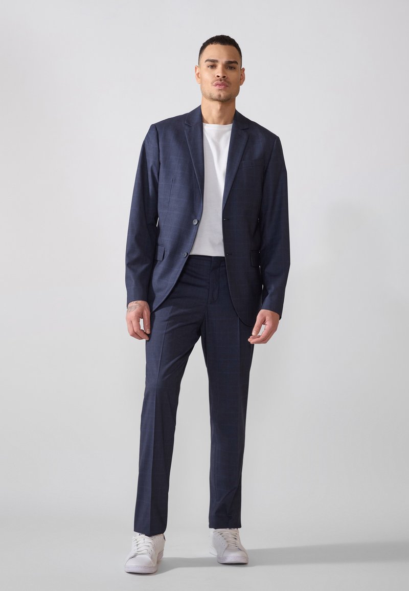 Costume à carreaux bleu marine avec une veste ajustée et un pantalon sur mesure, associé à un t-shirt blanc et des baskets blanches, texture de tissu lisse.