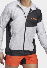 Męska kurtka TERREX w kolorach szarym i czarnym z ukośnym wzorem pasków, z pełnym zamkiem błyskawicznym oraz pomarańczowym logo na piersi. Posiada elastyczne mankiety.