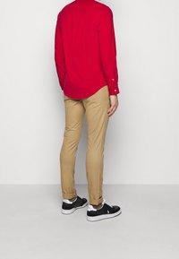 Maglietta rossa a maniche lunghe, pantaloni kaki slim fit e sneaker nere con accenti bianchi. La maglietta presenta polsini con bottoni e tessuto liscio.