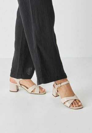 Beige sandaler med blokhæl, krydsede stropper og ankelspænde. Båret med sorte, strukturerede bukser med vide ben på en hvid overflade.