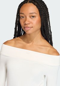 Biała bluzka bez ramiączek o gładkiej teksturze i subtelnym wytłoczonym logo na piersi. Dopasowany krój i długie rękawy.