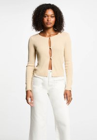 Beige ribbed cardigan with a keyhole front, long sleeves, and button closure. Paired with white wide-leg pants for contrast.