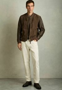 Jachetă bomber din suede maro și piele, cu manșete ribbate, asortată cu pantaloni crem și cizme negre. Design simplu și structurat.