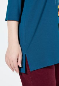 Túnica verde azulado con mangas tres cuartos que presenta aberturas laterales, combinada con pantalones burdeos. La tela parece suave y tiene una textura lisa.