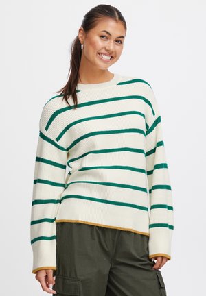 Maglione crema con righe orizzontali verdi, texture a coste, spalle scese e orlo giallo senape. Abbinato a pantaloni verde oliva.