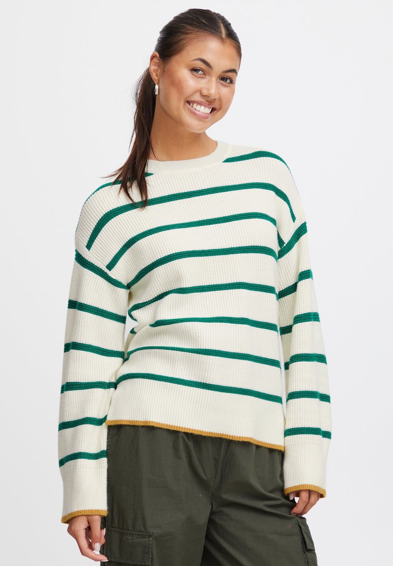 Maglione crema con righe orizzontali verdi, texture a coste, spalle scese e orlo giallo senape. Abbinato a pantaloni verde oliva.