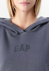 Heather grå hættetrøje lavet af blødt bomuldsstof med et mørkegrå "GAP" logo på forsiden, samt en hyggelig hætte foret med fleece.