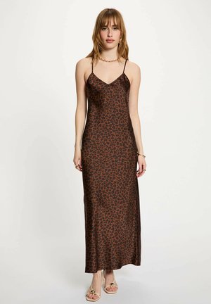 Giovane donna che indossa un vestito a sottoveste marrone e nero con stampa leopardata, con sottili spalline, accessori dorati e tacchi aperti beige, in piedi davanti a uno sfondo bianco.