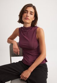 Burgundowa bezrękawnik z wysokim dekoltem, wykonany z gładkiego materiału. Modelka siedzi na metalowym krześle, ubrana w czarne szerokie spodnie.