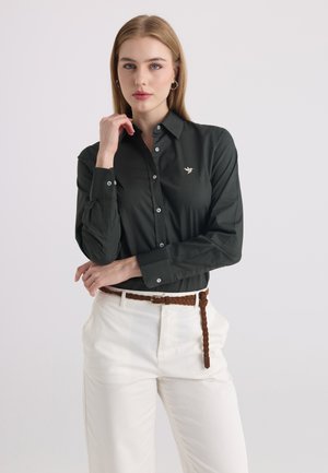 Donna con capelli biondi lisci indossa una camicia nera con bottoni e un piccolo logo di uccello e pantaloni bianchi con cintura marrone intrecciata.
