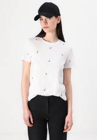 Witte katoenen T-shirt versierd met rode kersen borduursels, gecombineerd met zwarte broek. Het shirt heeft een klassieke ronde halslijn en korte mouwen.