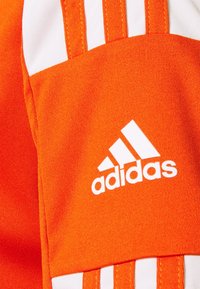 Oranžová sportovní bunda s bílými třemi pruhy a bílým logem Adidas. Materiál vypadá hladký s texturovaným povrchem.