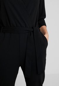 Mono negro con diseño envolvente, presenta un cinturón atado y bolsillos laterales. Confeccionado en una tela suave con un corte relajado y mangas de tres cuartos.