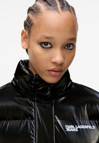Crna podstavljena jakna s visokim ovratnikom, s glossy finišom i bijelim logotipom "KARL LAGERFELD JEANS" na prsima.