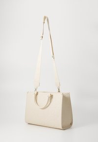 Sac fourre-tout beige avec un motif de logo embossé, deux anses supérieures et une bandoulière amovible. Texture lisse et forme structurée.